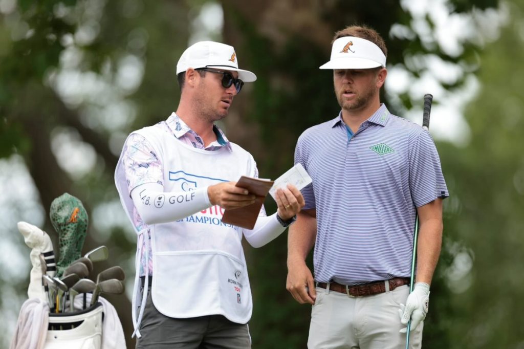 Two men discussing golf strategy outdoors, one holding a booklet, with a golf bag nearby.