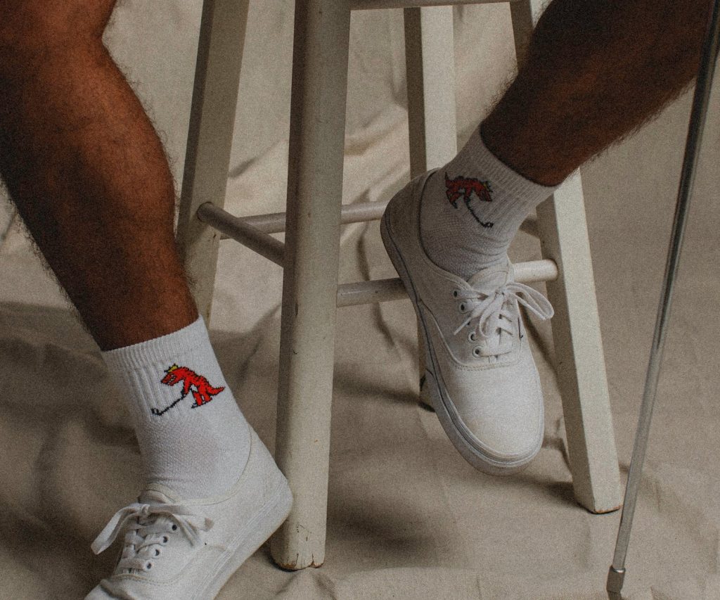 A close-up of a person's legs in white shoes and playful socks featuring a red dinosaur playing golf, near a white stool.