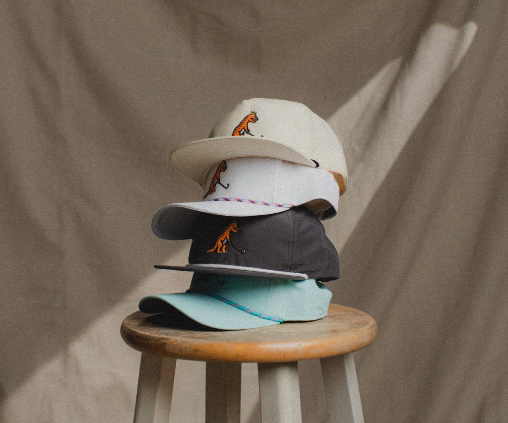 A stack of four colorful caps featuring a dinosaur emblem, displayed on a wooden stool against a neutral backdrop.