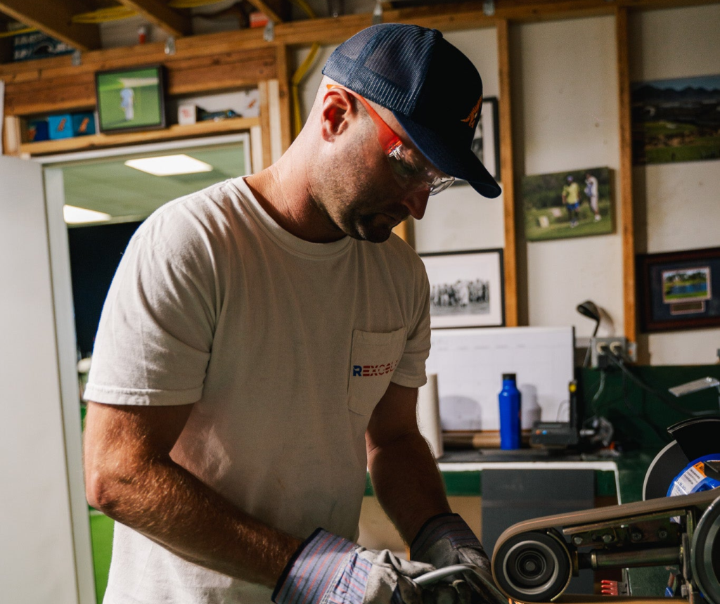 A man in a white shirt and cap is using machinery in a workshop, surrounded by golf-related images and equipment.