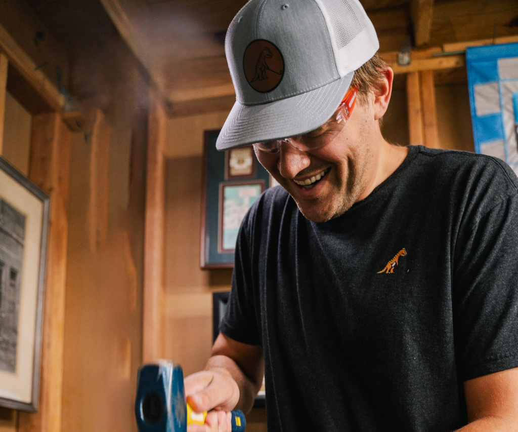 A person wears a gray cap and black shirt while using a tool in a wooden room filled with framed pictures and light steam.