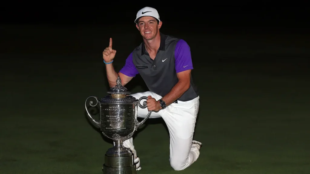 Rory McIlroy kneeling and smiling next to large golf trophy, pointing upward.