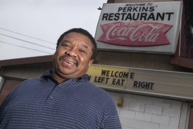 Man smiling in front of Perkins Restaurant sign with humorous message board