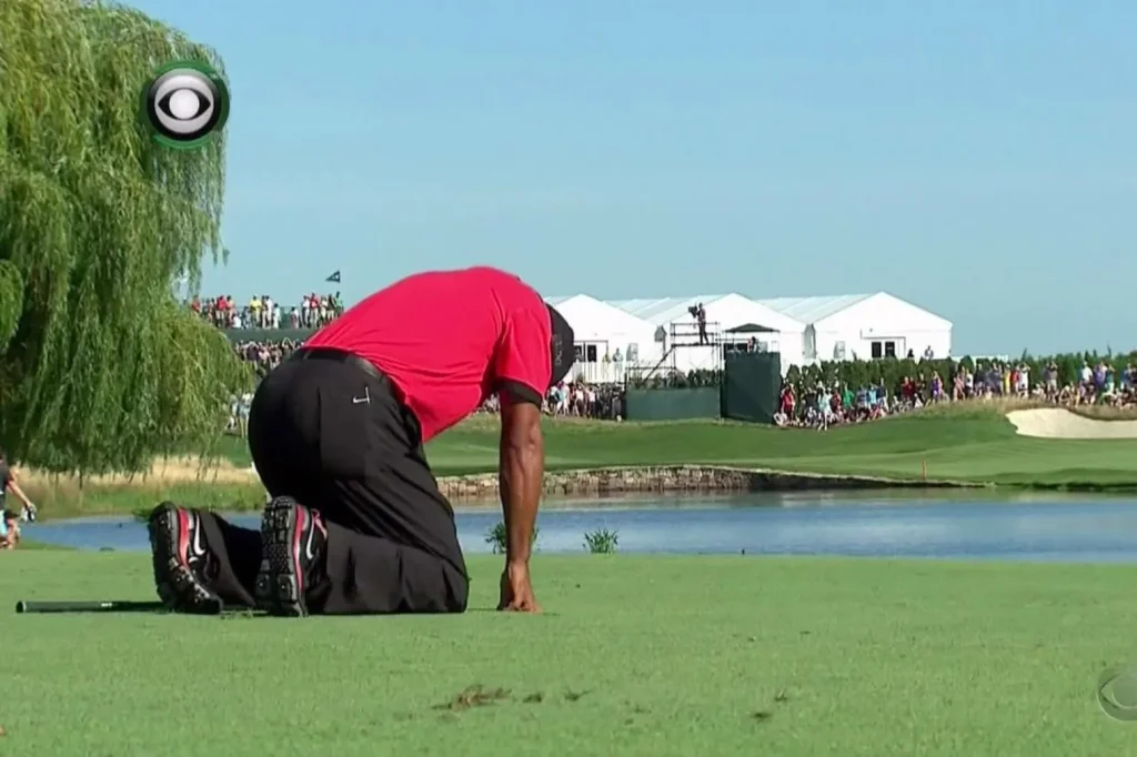 Tiger Woods kneeling on golf course in red shirt, appearing in pain or exhaustion.