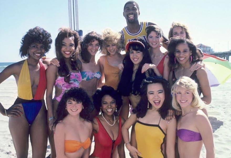 Magic Johnson smiling with group of women in swimsuits on beach.