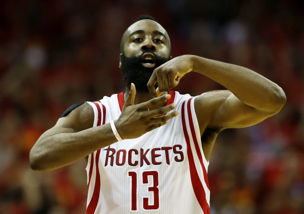 James Harden celebrating on court in Rockets jersey, gesturing with hand.