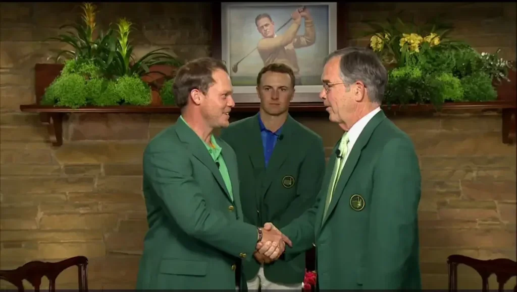 Jordan Spieth stands behind as Danny Willett shakes hands with Masters official indoors.