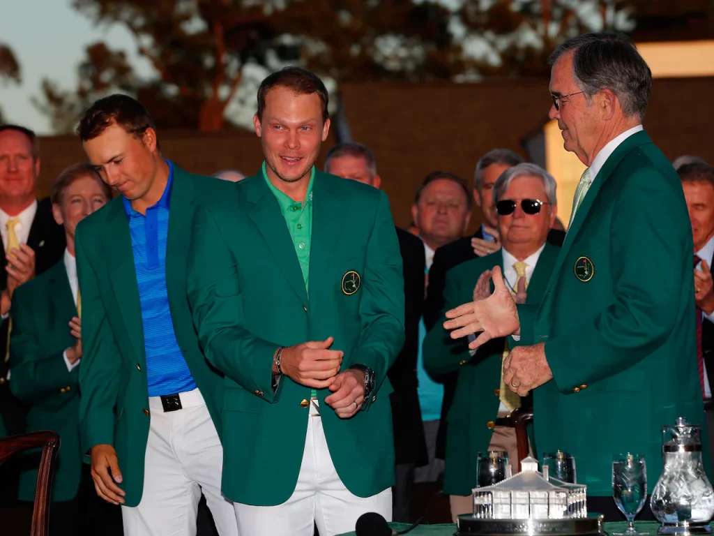 Jordan Spieth and Danny Willett wearing green jackets surrounded by crowd at ceremony.